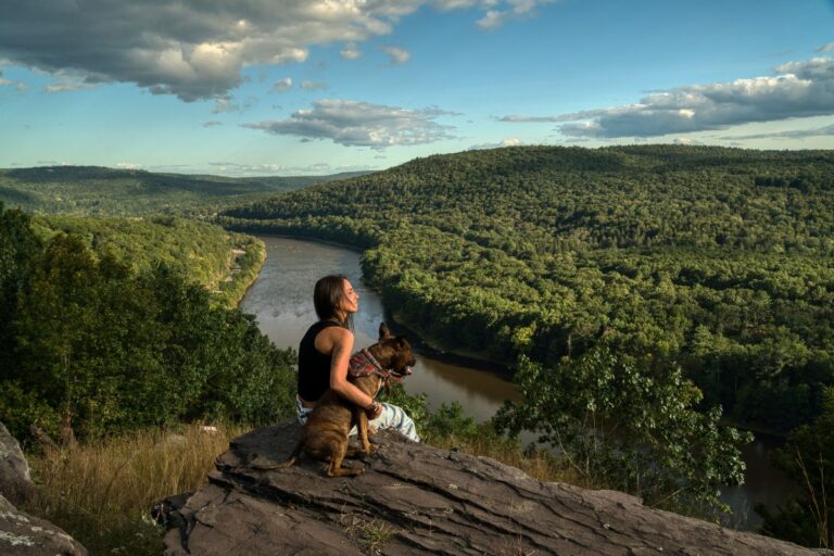 A woman sits on a rock with her dog, overlooking a scenic river valley surrounded by lush green hills under a partly cloudy sky.