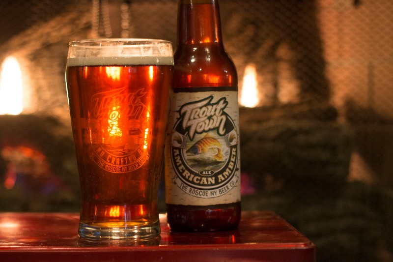 A close up of a bottle of Trout Town beer and a full glass of the beer in front of the fireplace. 